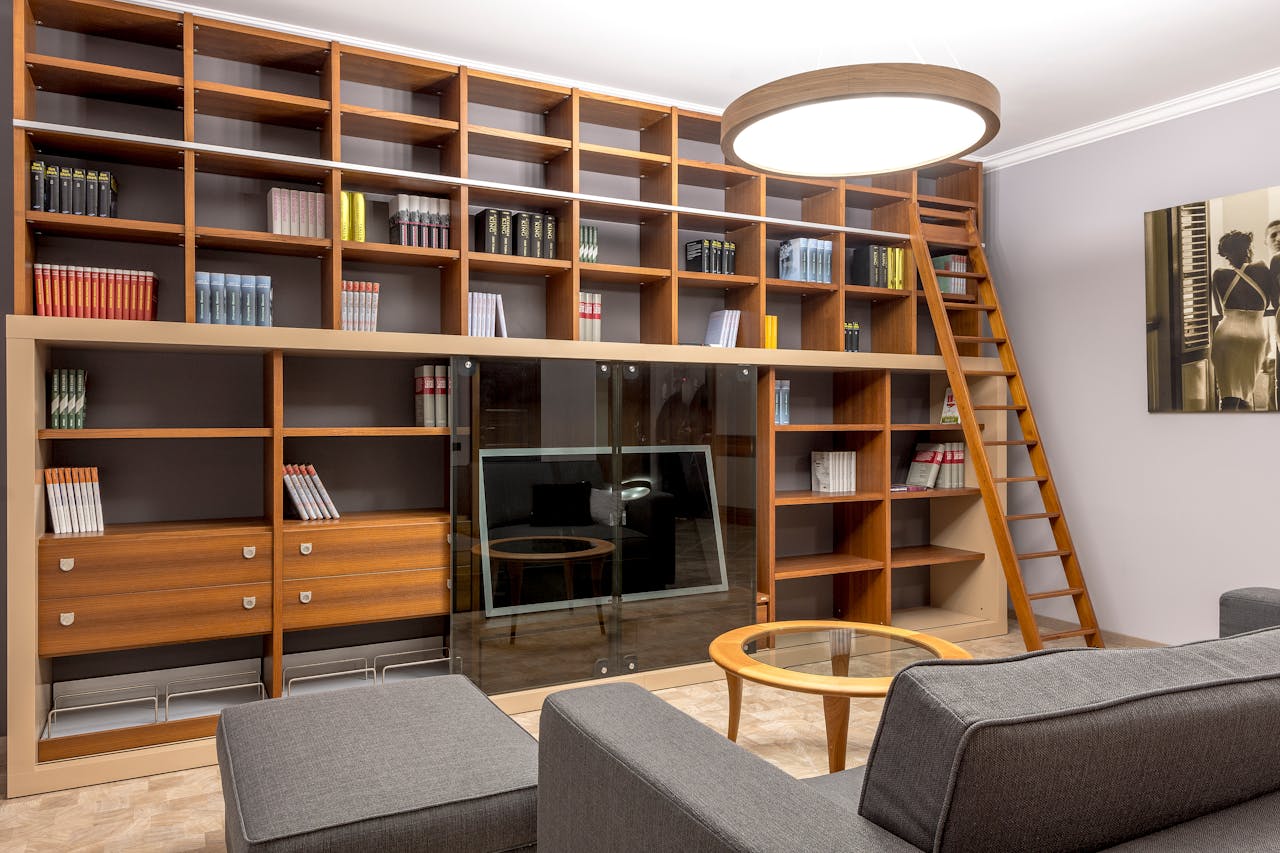 Stylish home library with wooden bookshelves, ladder, grey seating, and modern lighting.
